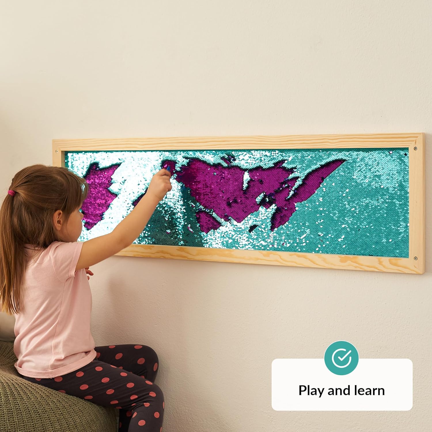Child interacting with a colorful sequins wall on a wall, with a 'Play and learn' label.